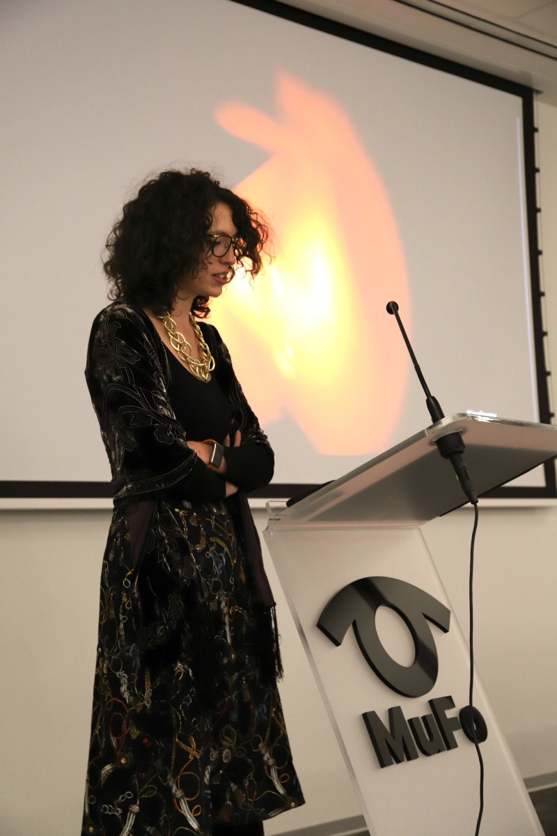 Młoda kobieta w ciemnej sukience przy mównicy, w tle ściana z ekranem